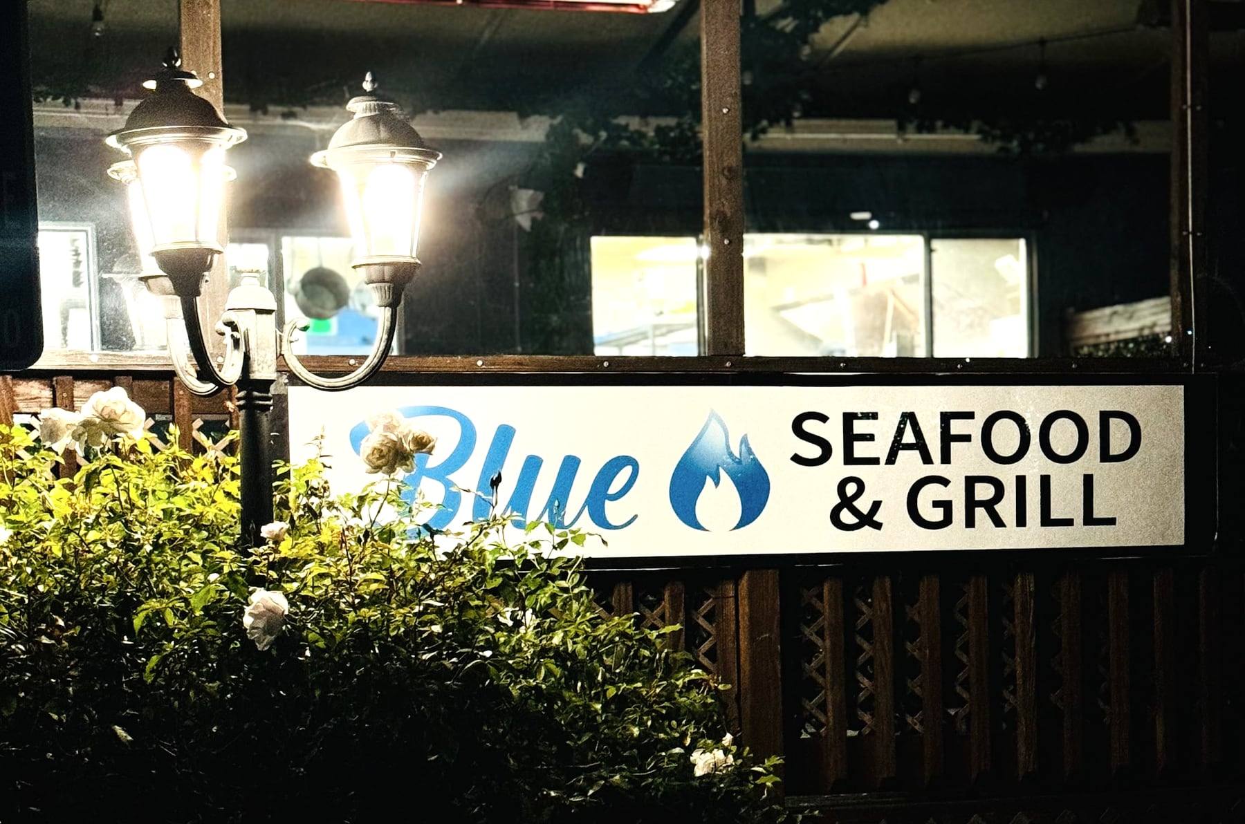 The Blue Seafood & Grill sign on the lattice fence at the back entrance at night, lit by a lamp post beside white roses