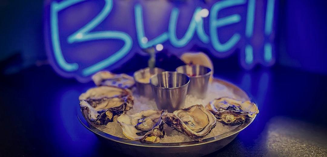 Oysters on the half shell on a platter of ice with sauce cups, in front of a glowing blue “BLUE” neon sign