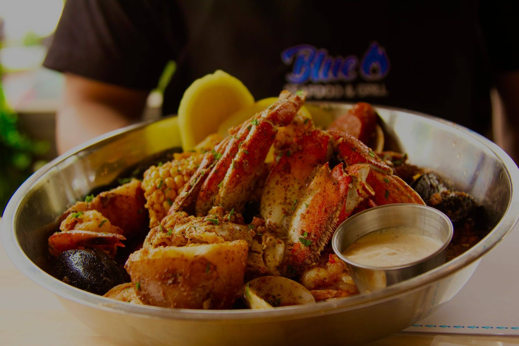 A steel bowl of seafood boil with crab legs, corn, mussels, clams and a cup of dipping sauce, held by a server in a Blue Seafood & Grill shirt