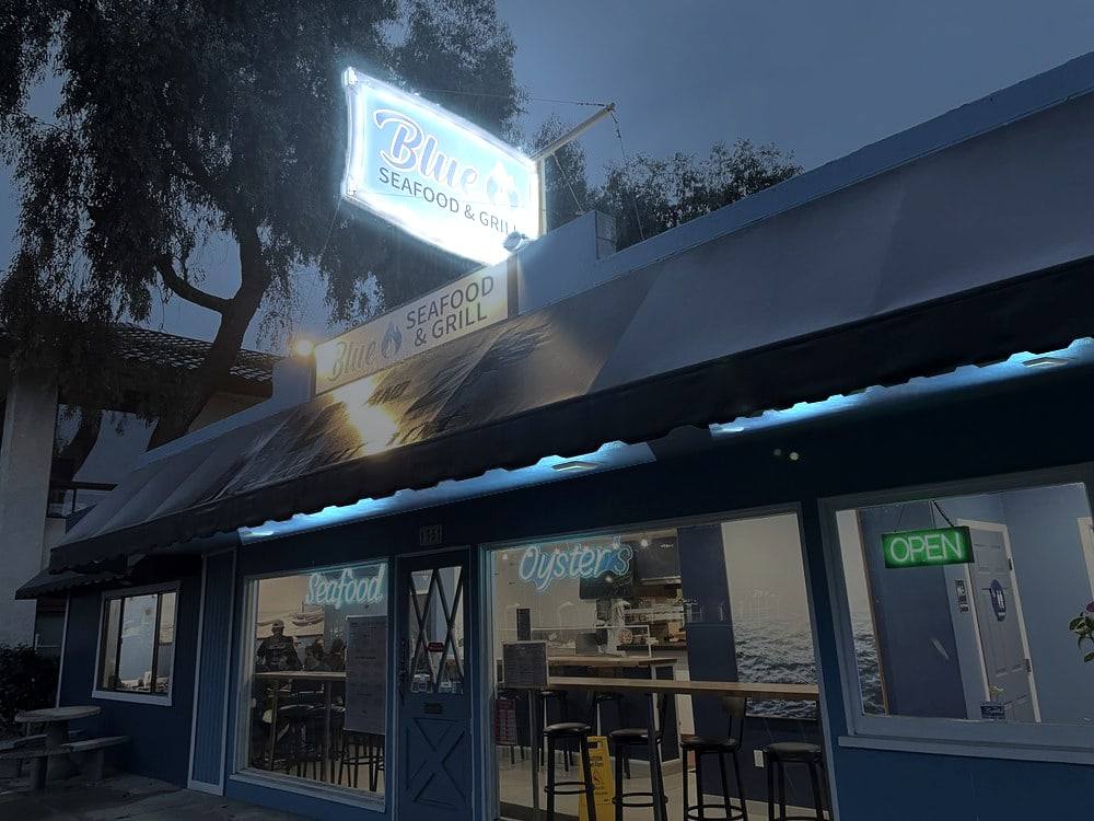 The Blue Seafood & Grill storefront at dusk: the lit blue sign above the awning and “Seafood” and “Oysters” neon in the windows